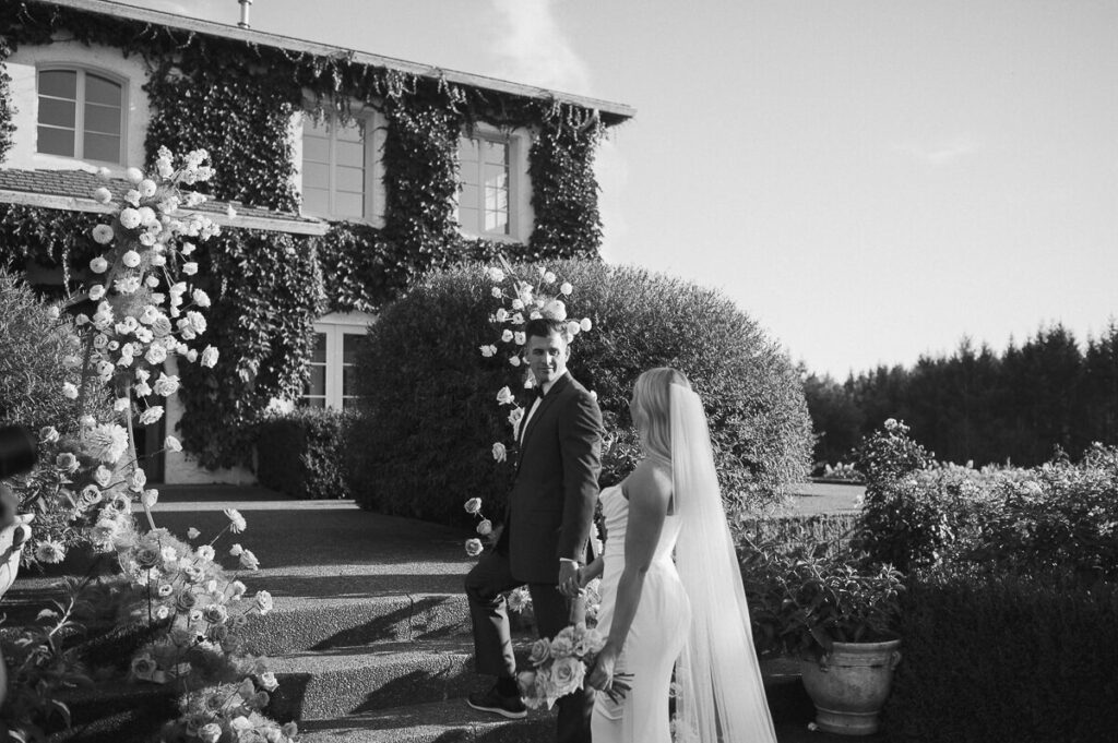 A bride and groom stand in front of their dahlia arch for their intimate garden wedding.